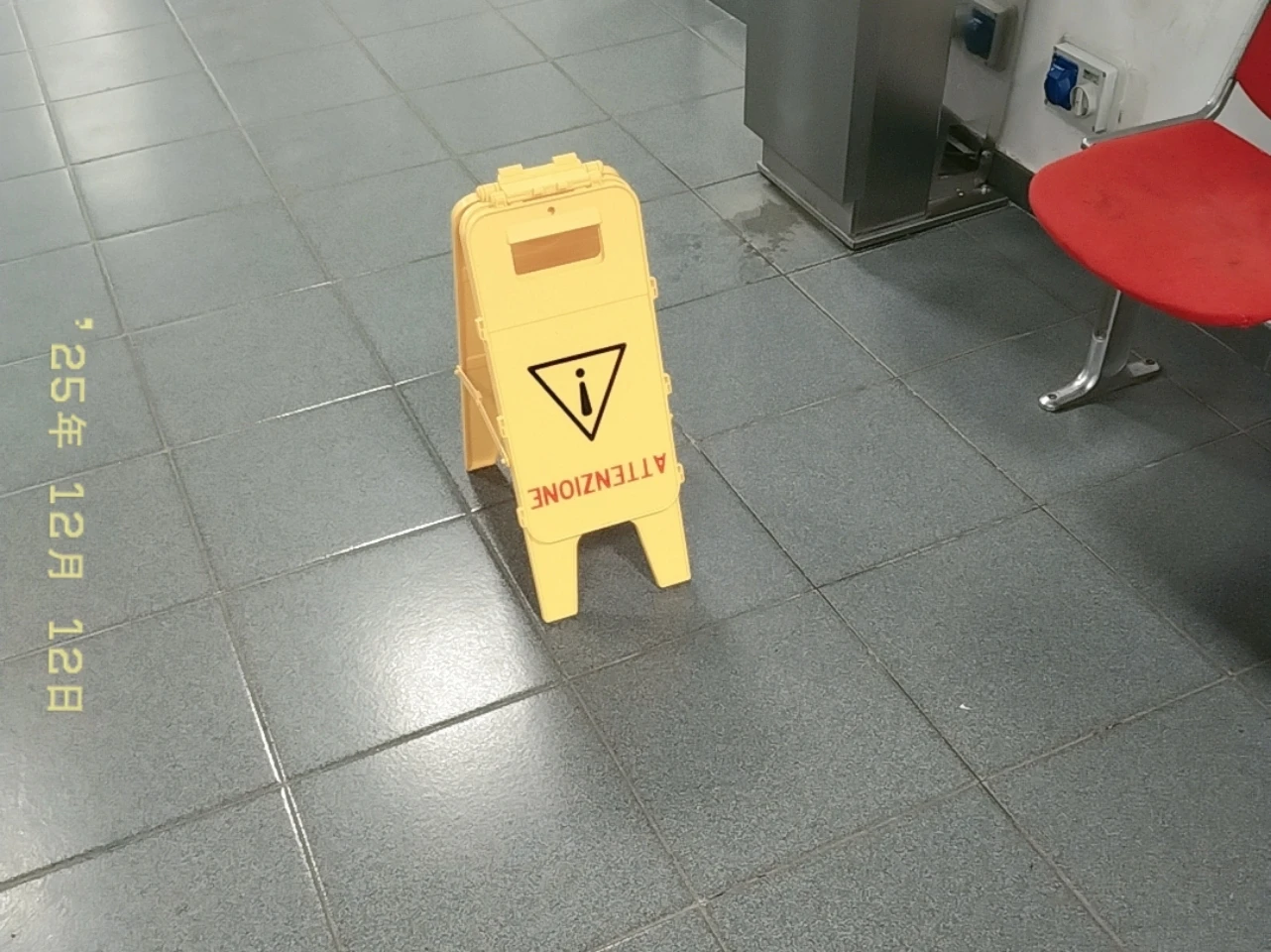 Cartello giallo di "ATTENZIONE" sul pavimento del corridoio di un edificio all'università, come descritto. Timestamp di oggi presente sulla foto.