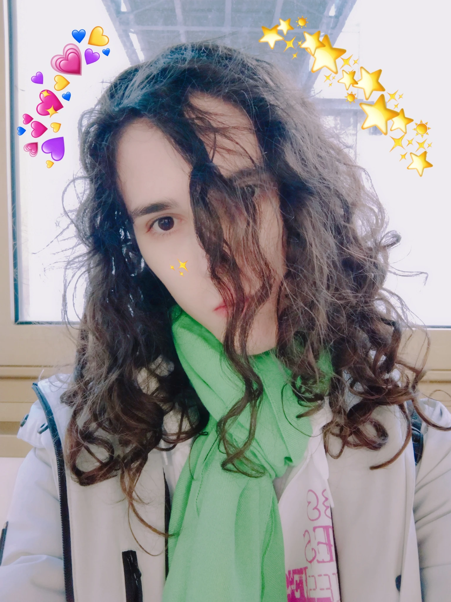 Selfie di spalle alla finestra sul lato interno del corridoio del secondo piano di F3 all'università, con la testa leggermente inclinata di lato, i capelli che un po' mi coprono la parte destra della faccia, e decorazioni di B612 di cuoricini e stelline accanto ai bordi della mia testa. Ho una sciarpa verde smeraldo, una felpa bianca con dei dettagli magenta che si intravedono, e un giubbino bianco-grigio.