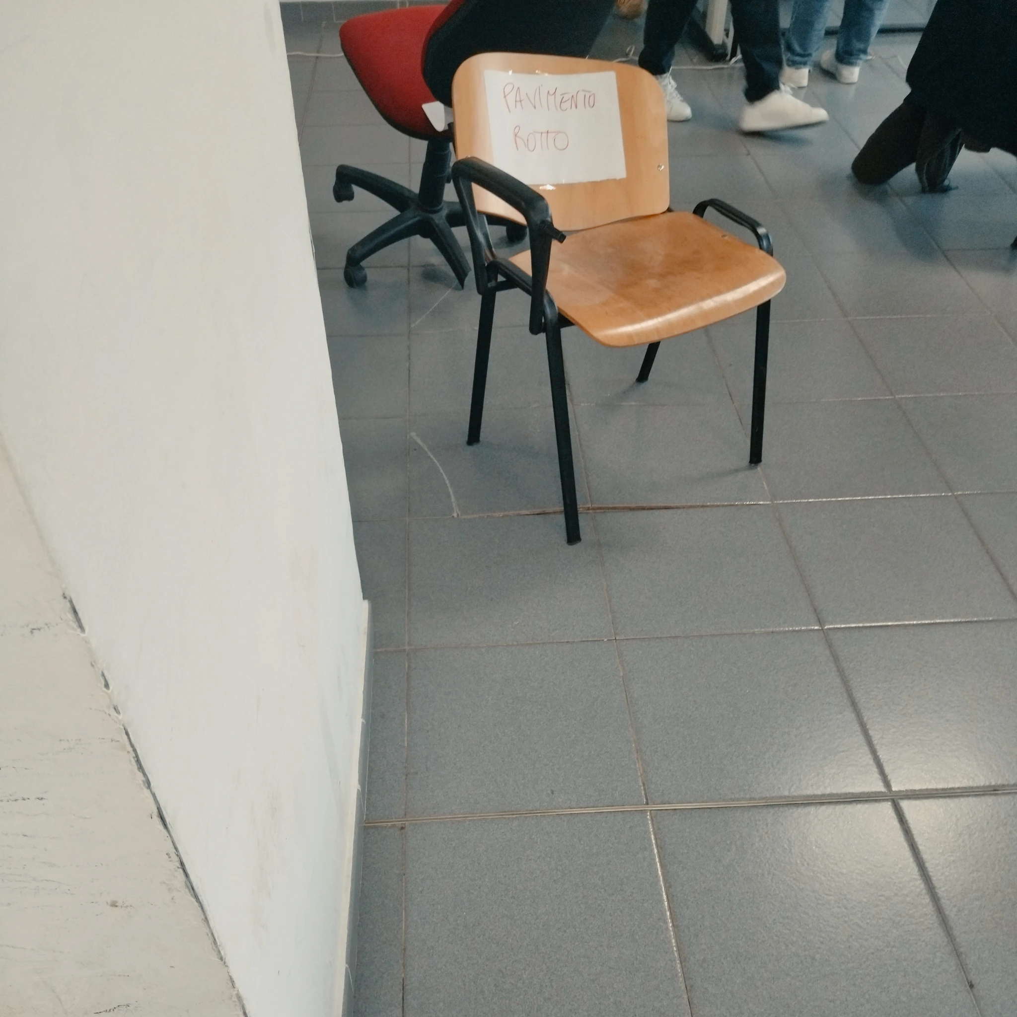 Foto dell'area come descritta. Pavimento a mattonelle quadrate di colore blu-grigio. Una sedie rossa e nera a ruote rotta che è quella in bilico dietro, e una sedia più classica di legno, a tavolino senza tavolino, e con lo schienale mezzo piegato davanti, con il cartello scritto in rosso:
PAVIMENTO
ROTTO