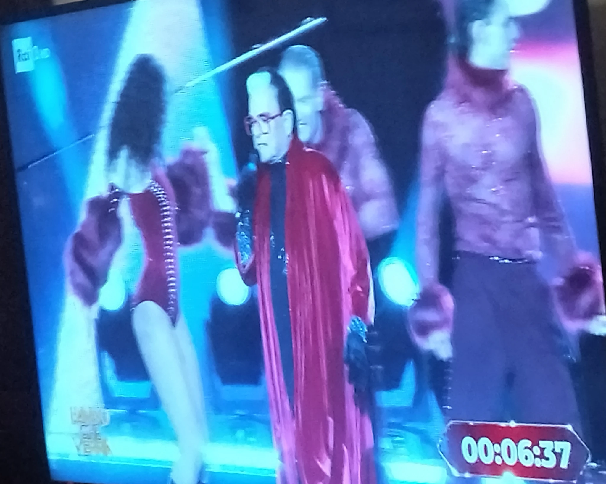 Foto fatta male, di sbieco, mezza inclinata, alla TV su un frame della trasmissione, a minuto 00:06:37, che mostra 2 tizi e 1 tizia che ballano, e Malgioglio davanti.